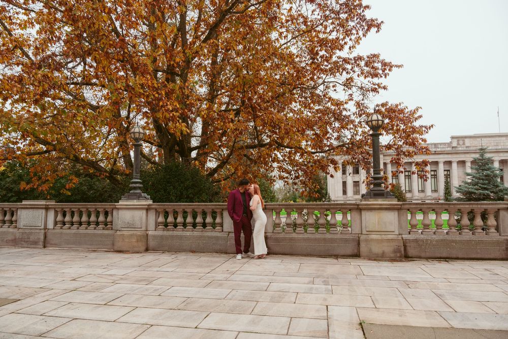 A couple stands on a stone terrace overlooking an autumn tree with orange foliage and a historic building in the background.