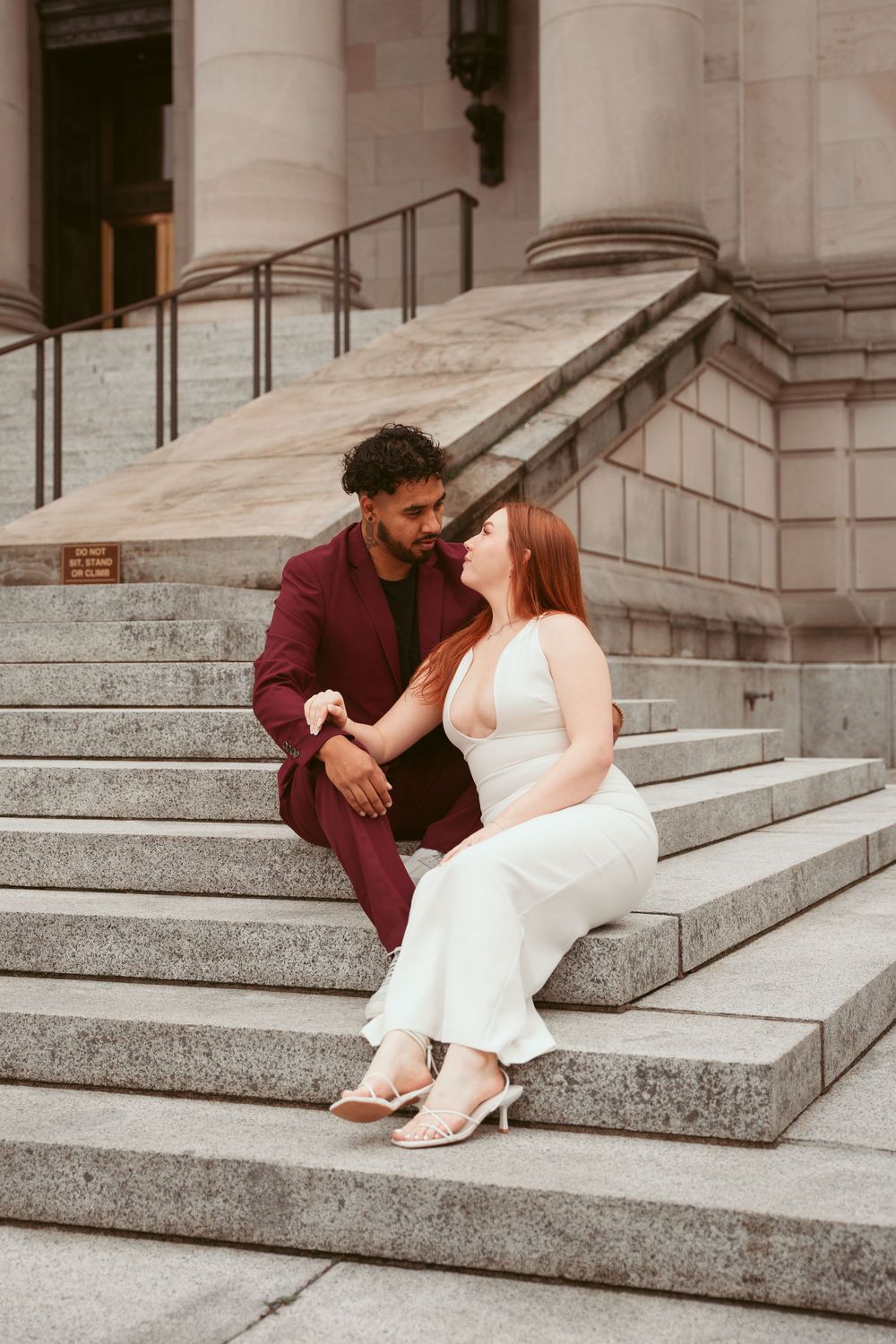 A couple in wedding attire shares an intimate moment while sitting on stone steps of a grand building.