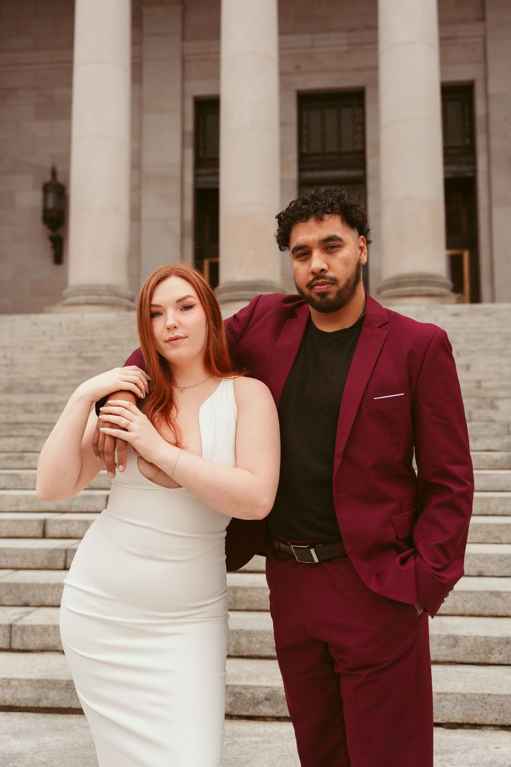 Elegant couple poses on courthouse steps with bride in white dress and groom in burgundy suit.