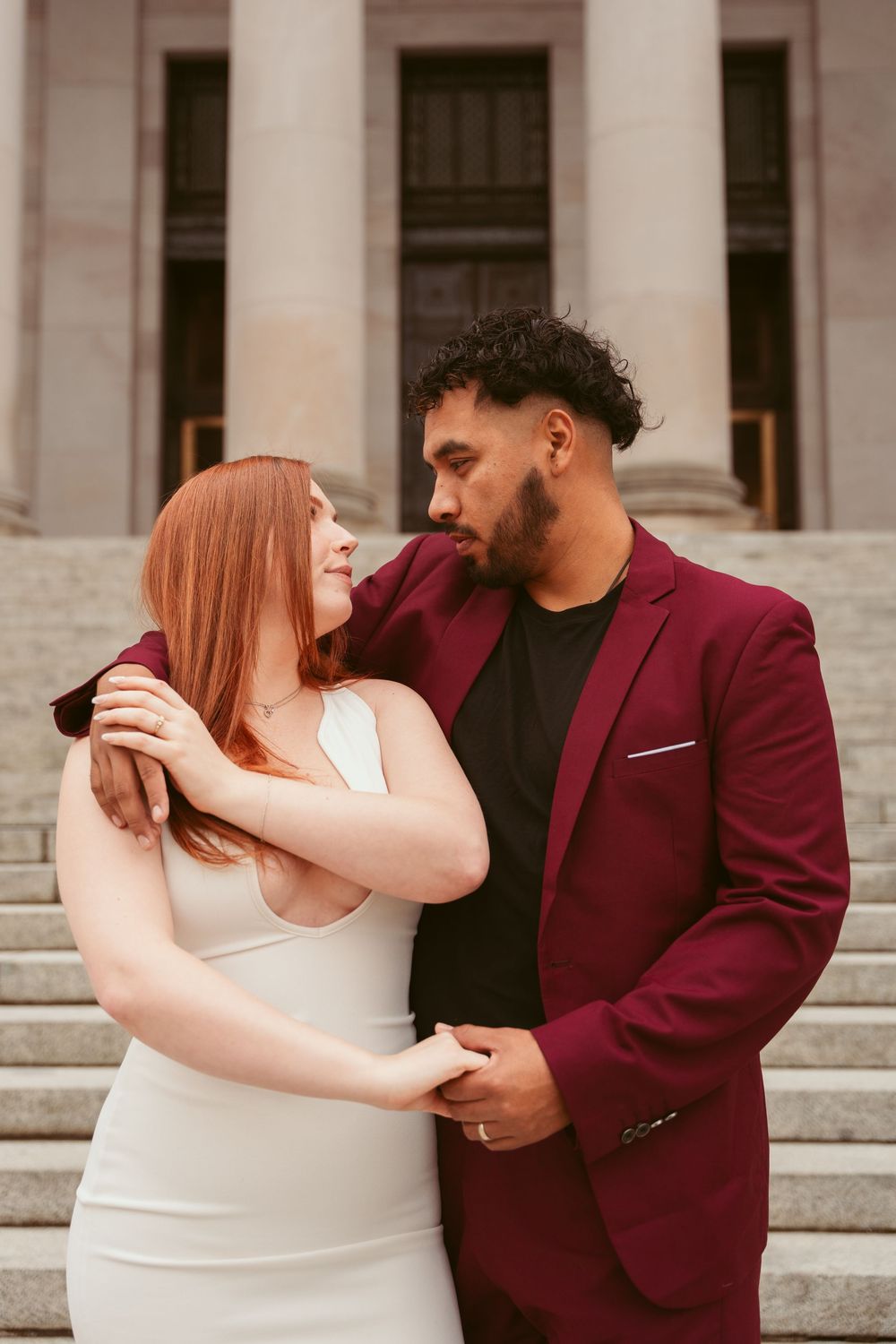 A romantic couple embraces on stone steps, with the person in a burgundy blazer and partner in white dress gazing at each other.