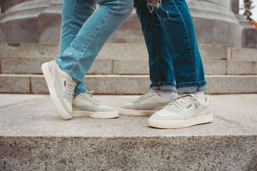 Two people wearing blue jeans and white sneakers stand facing each other on concrete steps.