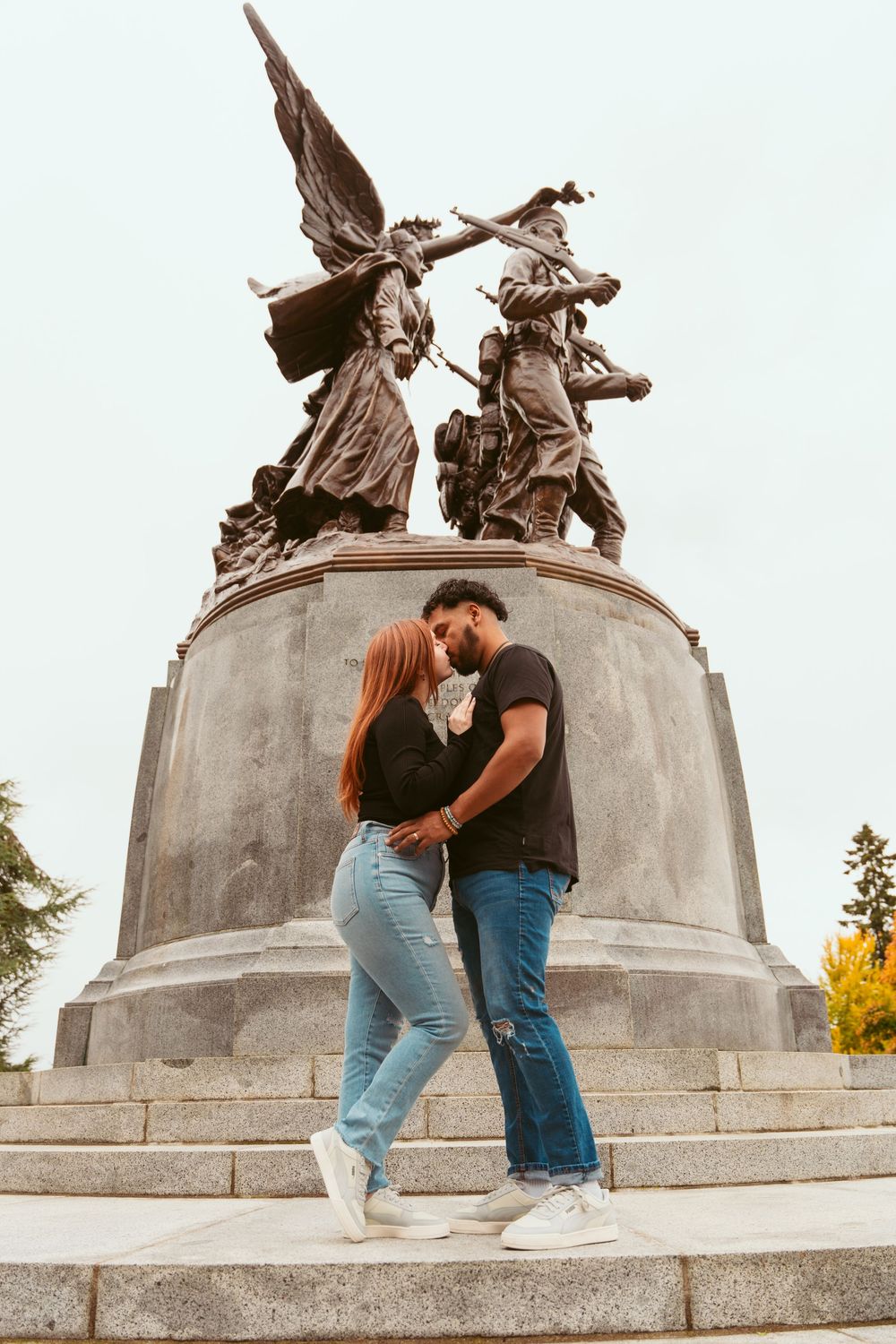 Couple embraces on stone steps in front of dramatic winged statue monument at golden hour.