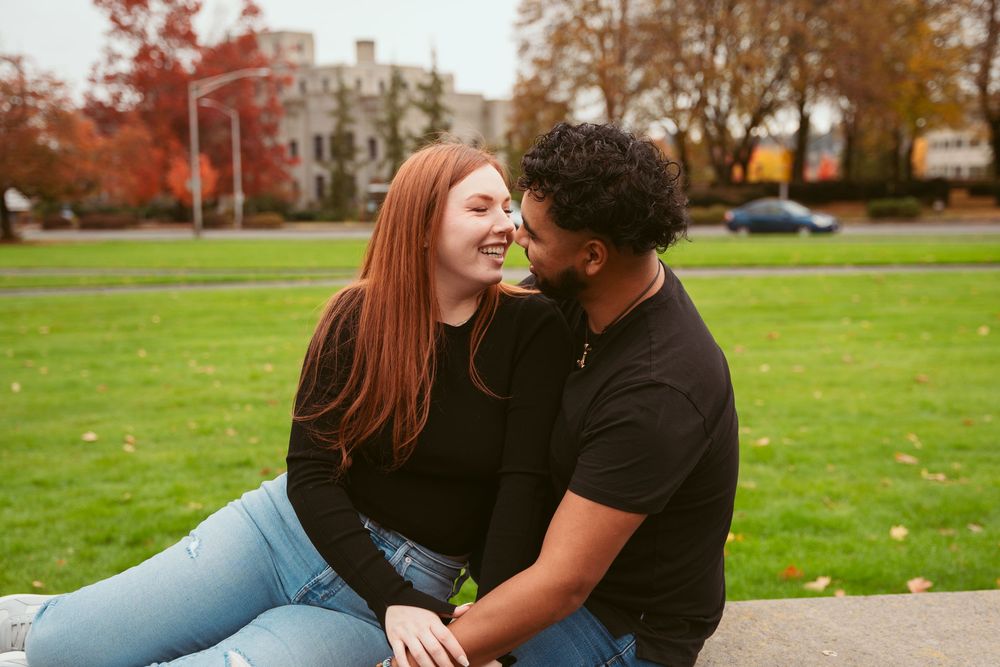 A joyful couple sits together laughing on grass with autumn trees and buildings in the background.