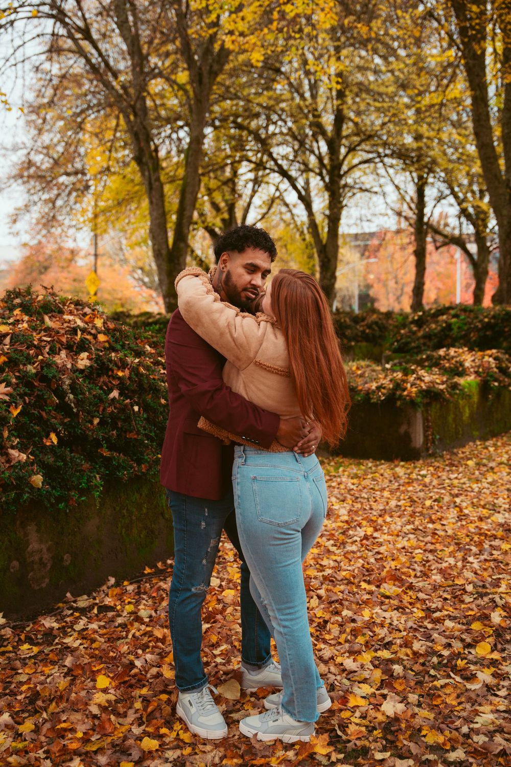 A couple embraces on an autumn path covered in fallen yellow leaves next to a peaceful stream.