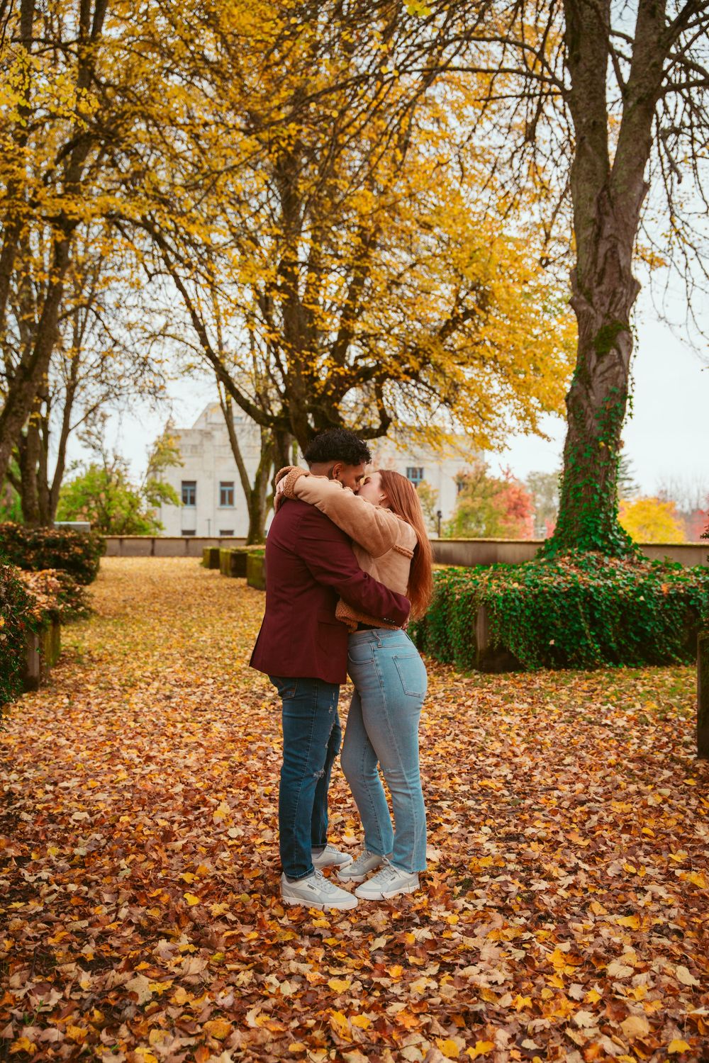 Couple embracing on fallen autumn leaves with white building in background.