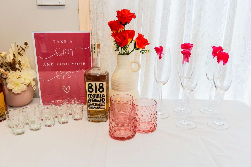 Pink and red wedding decor with roses in glass vases, vintage glassware, and bar supplies on a white tablecloth.