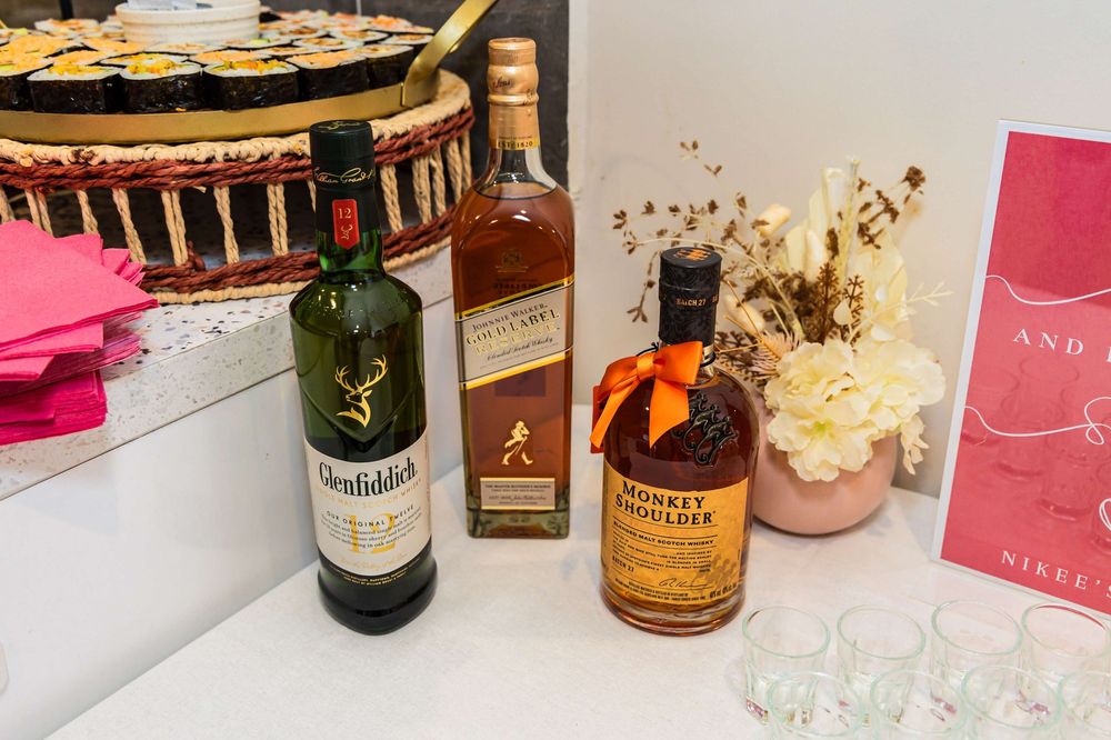 Wedding bar setup featuring whiskey, wine bottles, pink signage and floral arrangements on a white tablecloth.