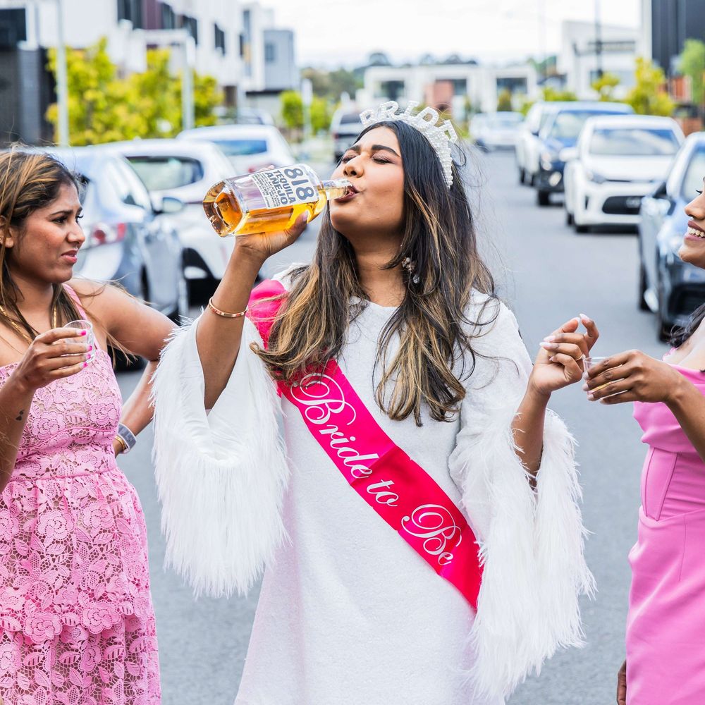 Bride in white dress and Bride Squad in pink dresses drinking champagne during outdoor celebration.