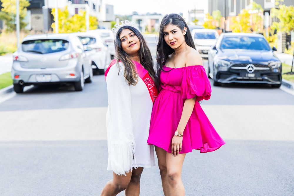 Two women pose together on a city street wearing pink and white outfits with parked cars in the background.