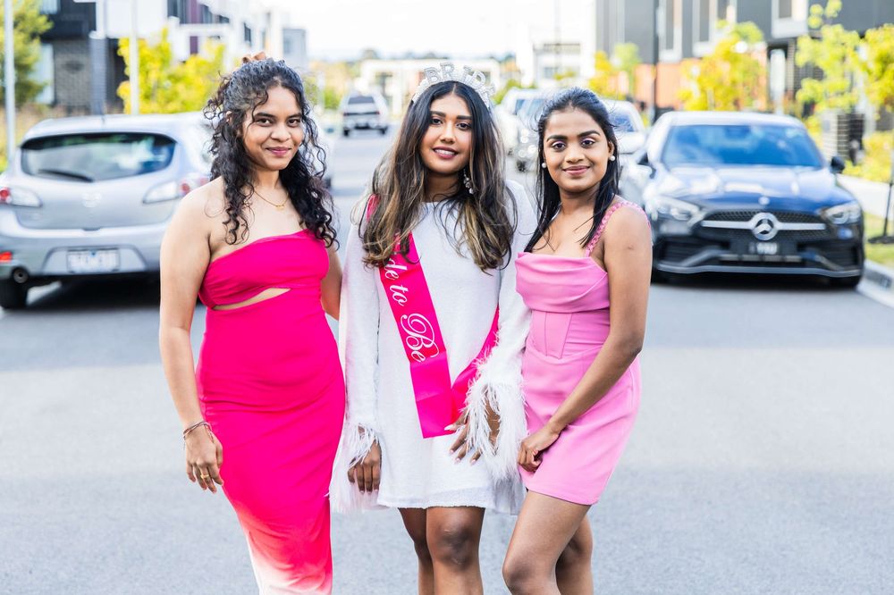 Three friends in bright pink and white dresses pose together on a city street with parked cars in the background.