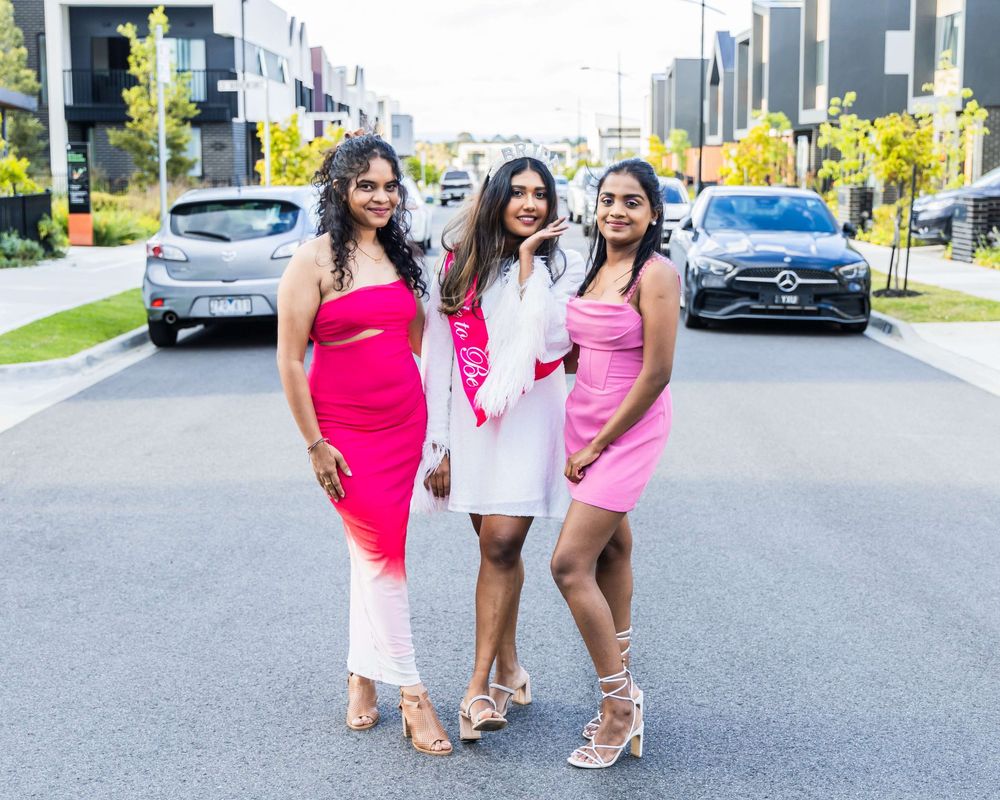 Three people pose together wearing pink and white dresses standing on a street with luxury cars in the background.