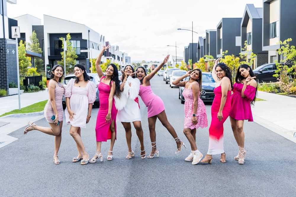 Group of friends wearing pink and white dresses pose joyfully together on a modern residential street.