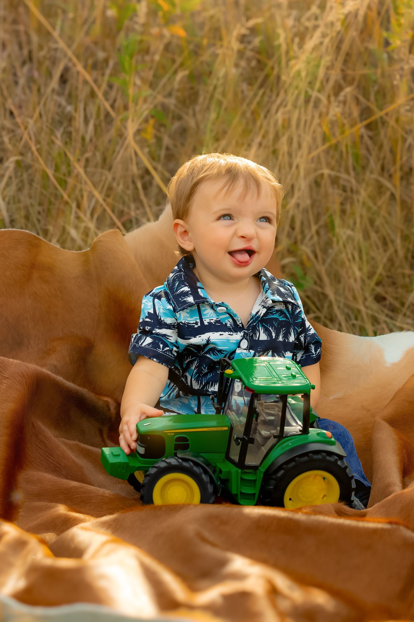 A happy toddler playing with a green John Deere toy tractor on a brown blanket outdoors.