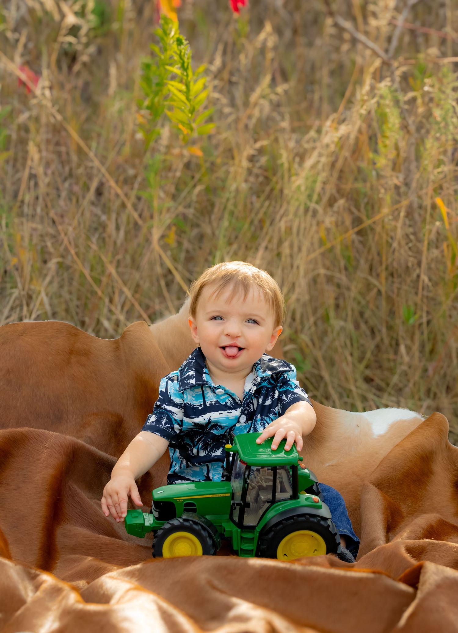 Toddler in blue playing with green tractor toy on brown blanket with red fall leaves in background.