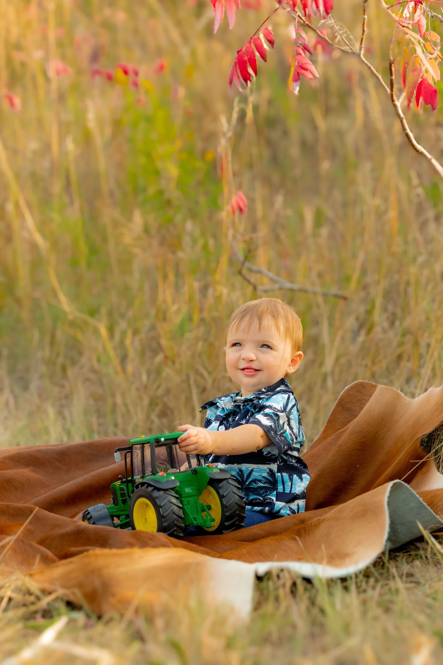 Child lying on brown blanket with toy tractor against autumn foliage background.