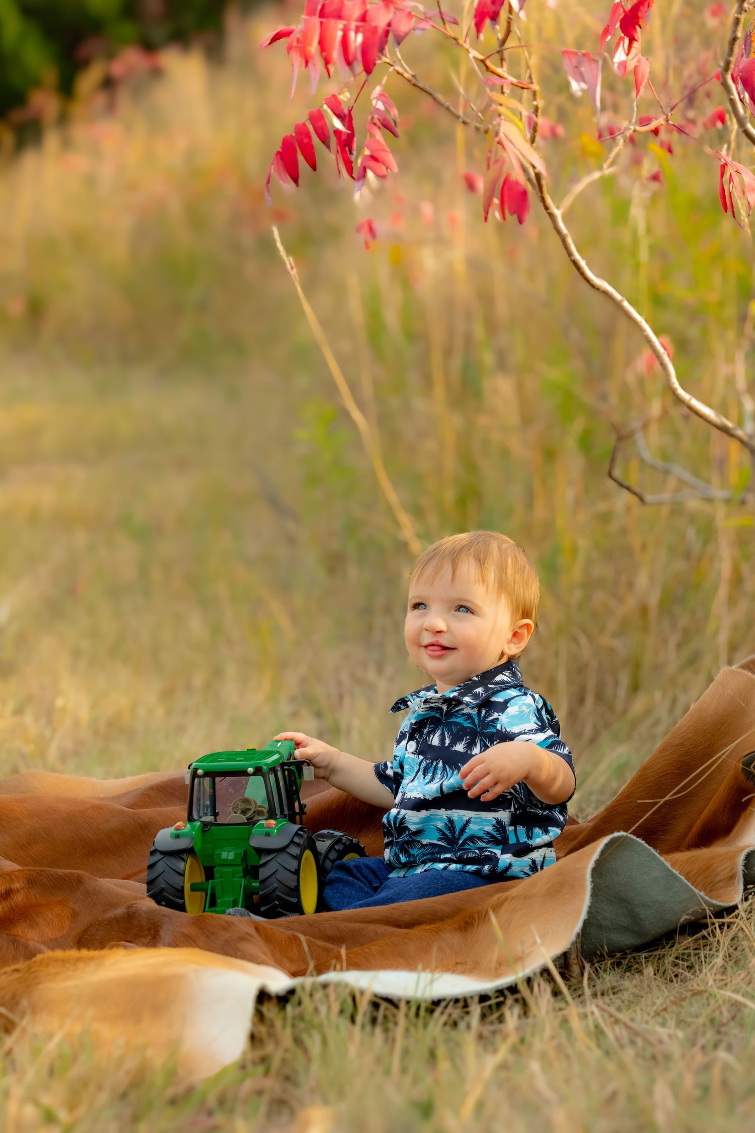 Child playing with toy tractor while lying on brown blanket outdoors with red autumn foliage.