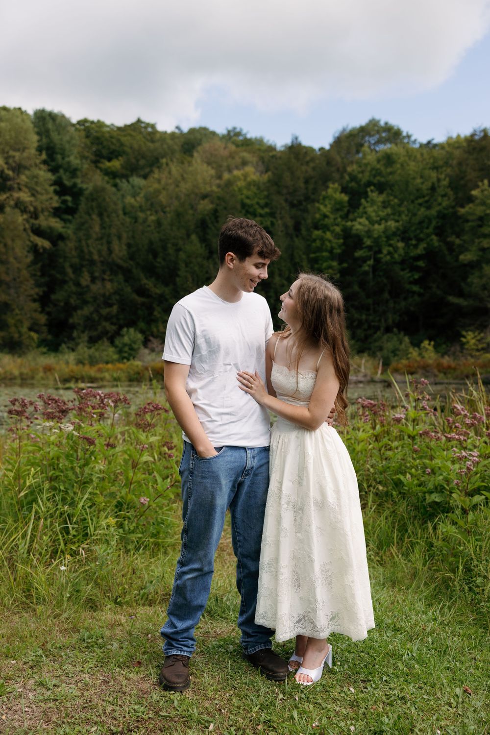 Young couple in white dress and jeans smiling at each other in a lush green outdoor setting.
