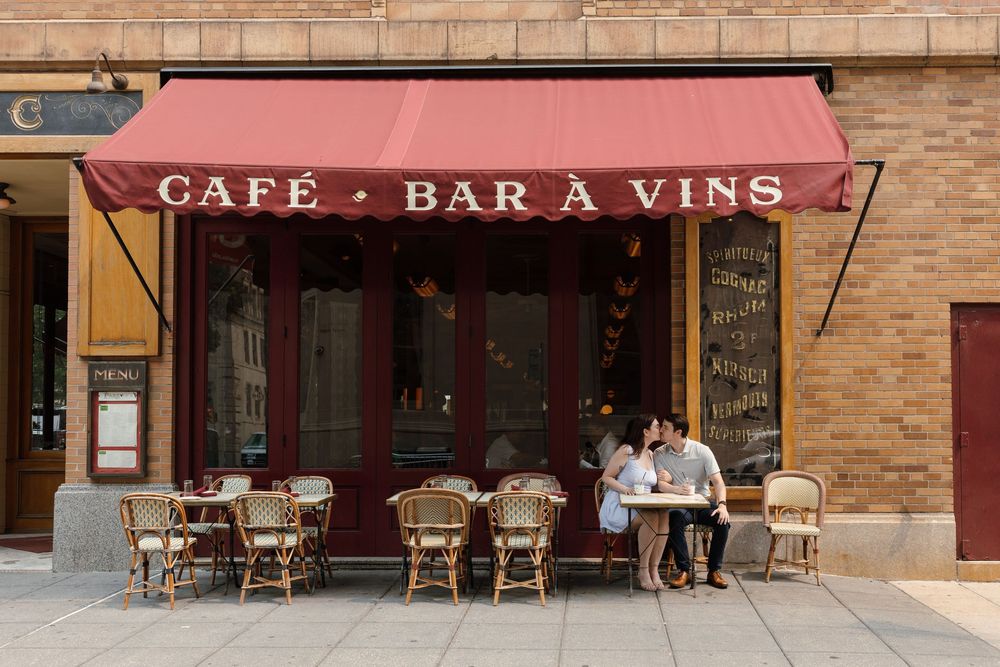 Couple sitting at outdoor cafe table under red awning reading "Café Bar à Vins" on a sunny day.