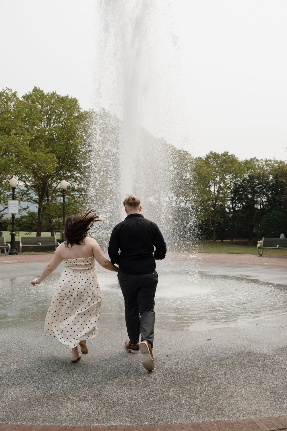 Two people run joyfully through a water fountain spray in a park on a sunny day.