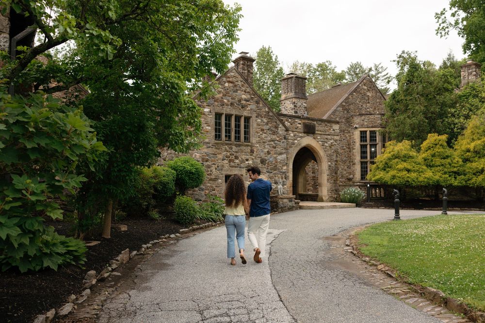 Couple walking hand-in-hand toward a historic stone manor surrounded by lush green trees on a paved pathway.