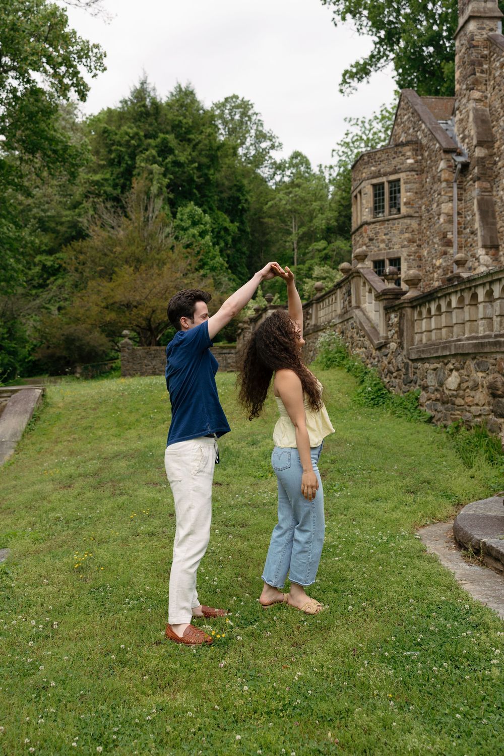 A couple dances on a green lawn beside a stone castle surrounded by lush trees.