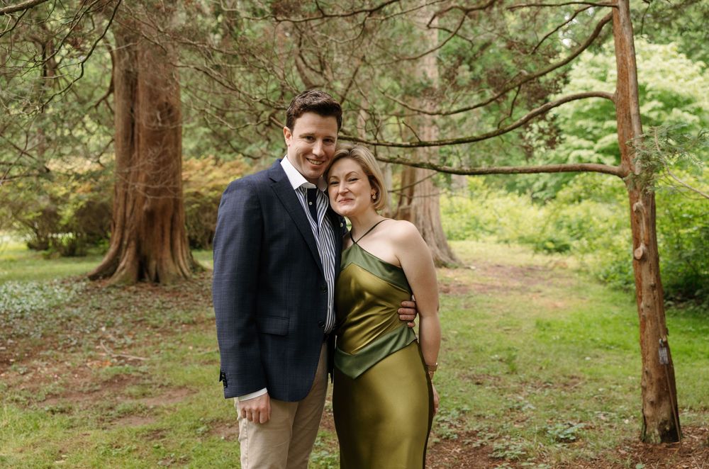 Couple posing outdoors in formal attire, man in navy suit, woman in gold dress, surrounded by tall trees.