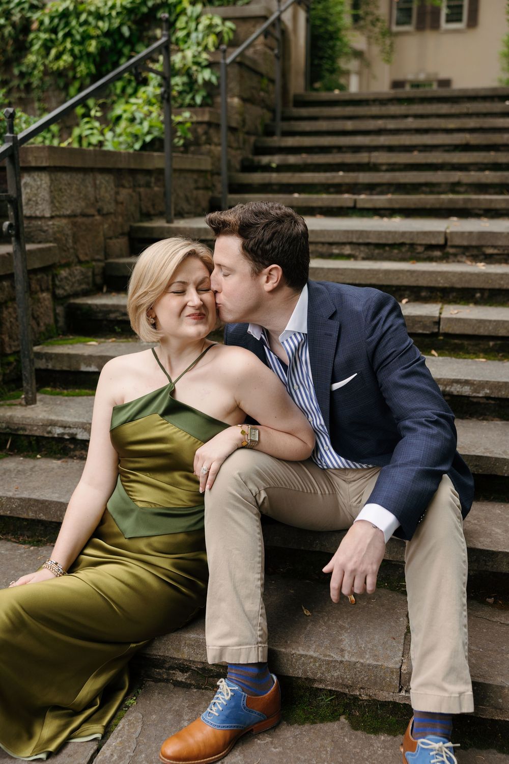 A couple sits on stone steps, man in navy suit kissing woman in olive satin dress, surrounded by lush greenery.