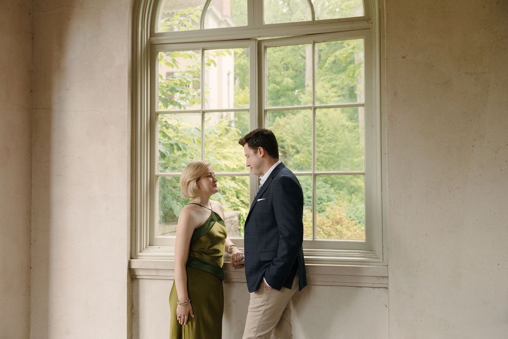 Couple in elegant attire standing by a large window with green foliage, woman in olive dress, man in navy blazer.