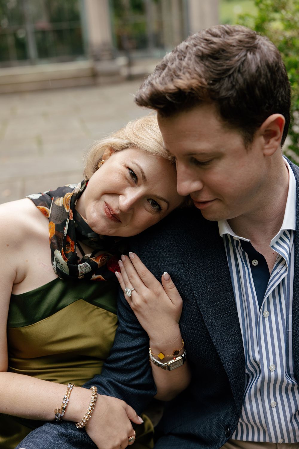 A couple shares a tender moment outdoors, woman in green dress resting head on man's shoulder, wearing engagement ring.