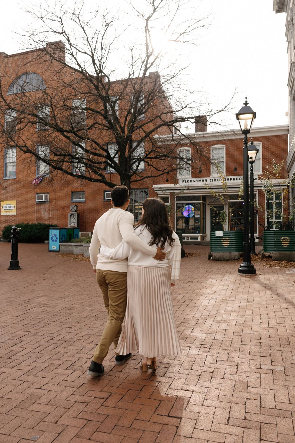 Two people walk along a brick courtyard surrounded by historic brick buildings and lamp posts on an overcast day.