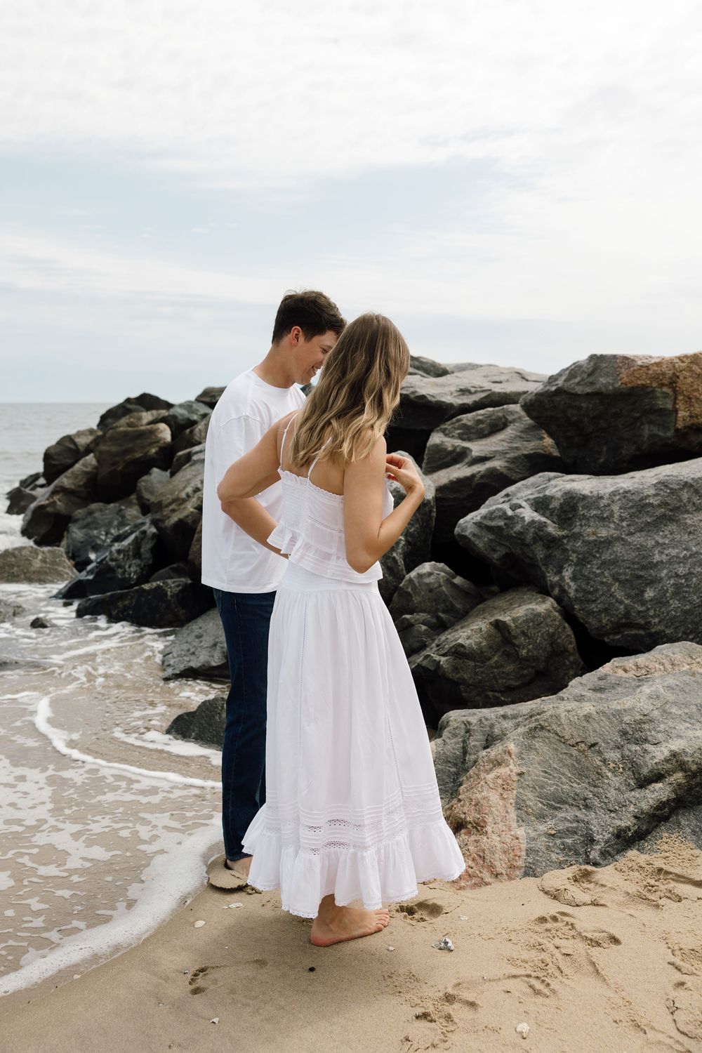 Couple in white outfits standing together on rocky beach coastline.