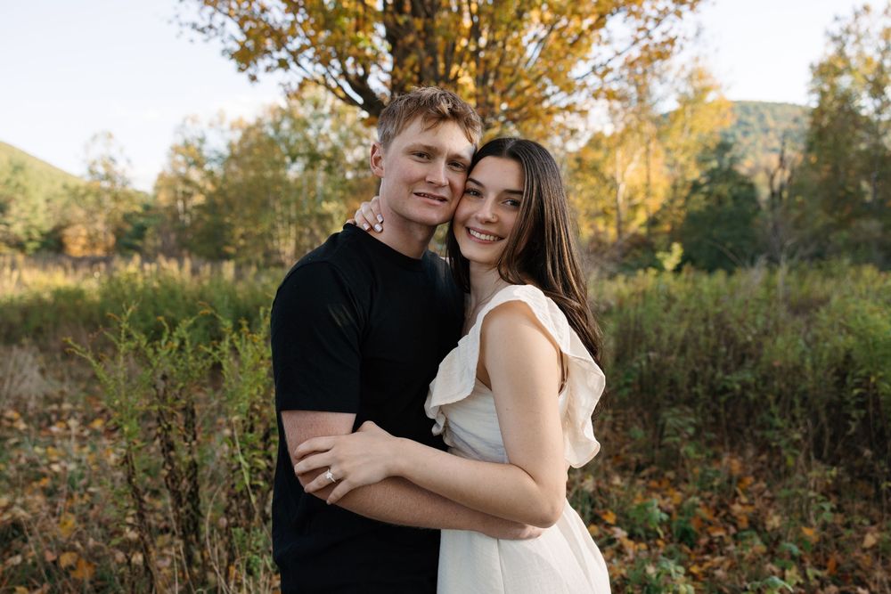 A couple poses together in a romantic outdoor setting during golden hour with autumn foliage in the background.