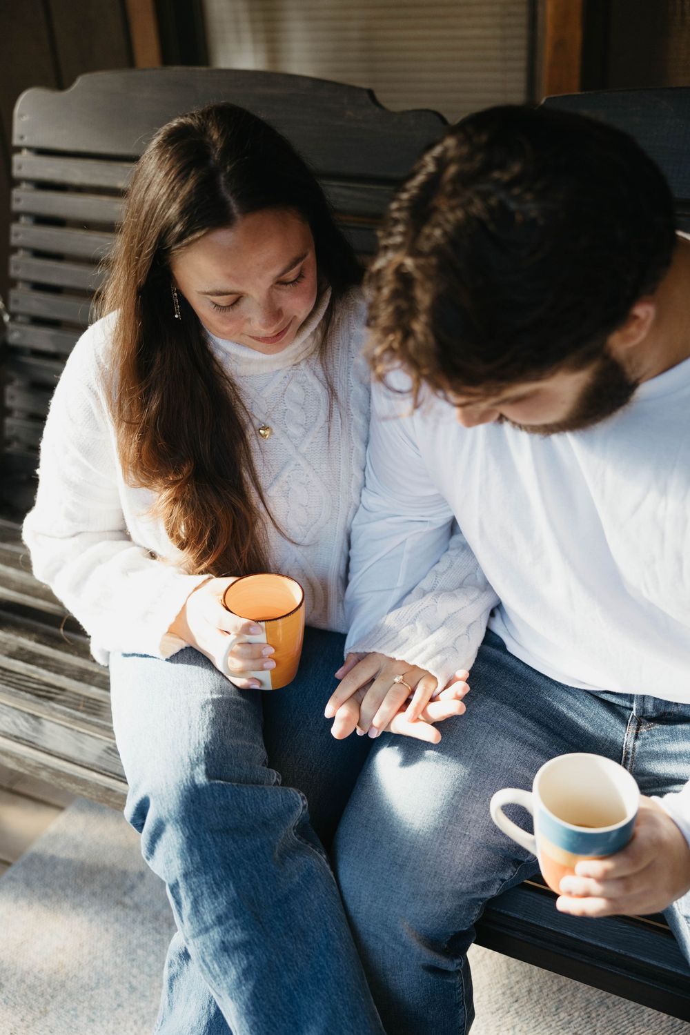 Two people in casual attire sit close together on a porch sharing coffee and intimate conversation.