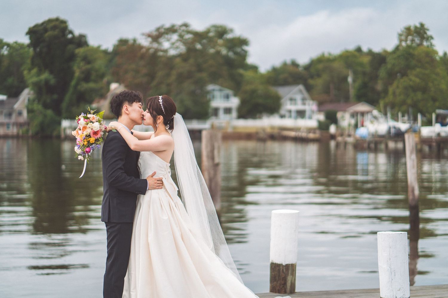 Newlyweds share an intimate moment on a dock by the water with houses in the background.