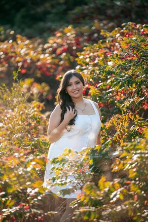 A person in a white top poses among vibrant autumn foliage with golden sunlight filtering through the leaves.