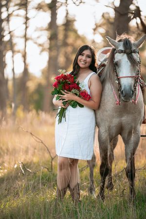 A white dress-clad figure holds red roses while standing beside a gray horse in a sunlit autumn field.