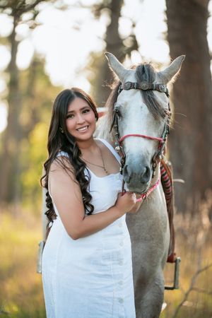 A white horse poses with someone in a white dress during an outdoor photoshoot among pine trees.