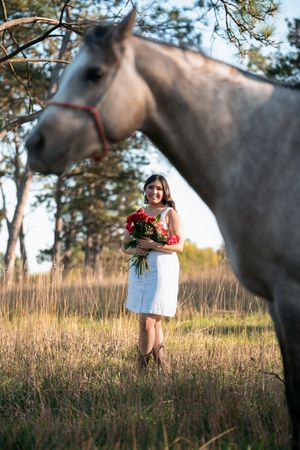 A figure in a white dress holds red roses while standing in a grassy field with a gray horse in the background.