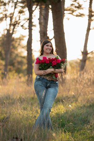 Person in jeans holds vibrant red roses while standing in a sunlit field with trees at golden hour.