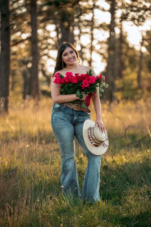 Person in jeans holding red roses and a cowboy hat poses in a golden sunlit field at sunset.
