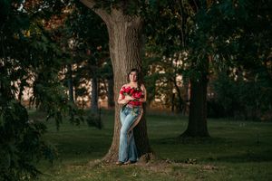 A person in a red top and blue skirt poses against a large tree trunk in a serene wooded setting at sunset.