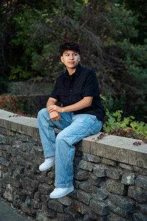 Person in black shirt and blue jeans sits casually on stone wall surrounded by greenery in outdoor setting.