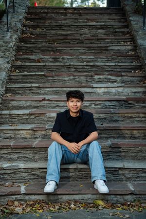 A person sits on weathered wooden steps against a rustic wall wearing a black shirt and blue jeans.