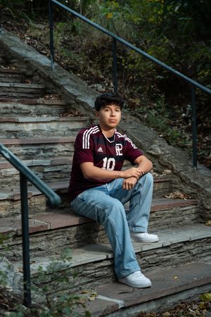 Student athlete in maroon sports jersey and jeans sits thoughtfully on outdoor wooden steps.