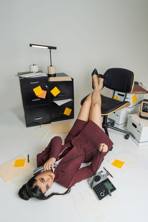 Businesswoman in burgundy suit lying on office floor talking on phone, surrounded by papers and sticky notes.