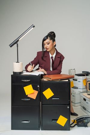 Businesswoman in burgundy suit writing notes at black filing cabinets with sticky notes and desk lamp in office.