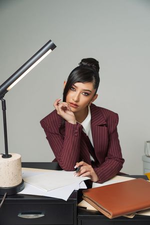 Young professional woman in striped blazer sitting thoughtfully at office desk with lamp and notebooks.