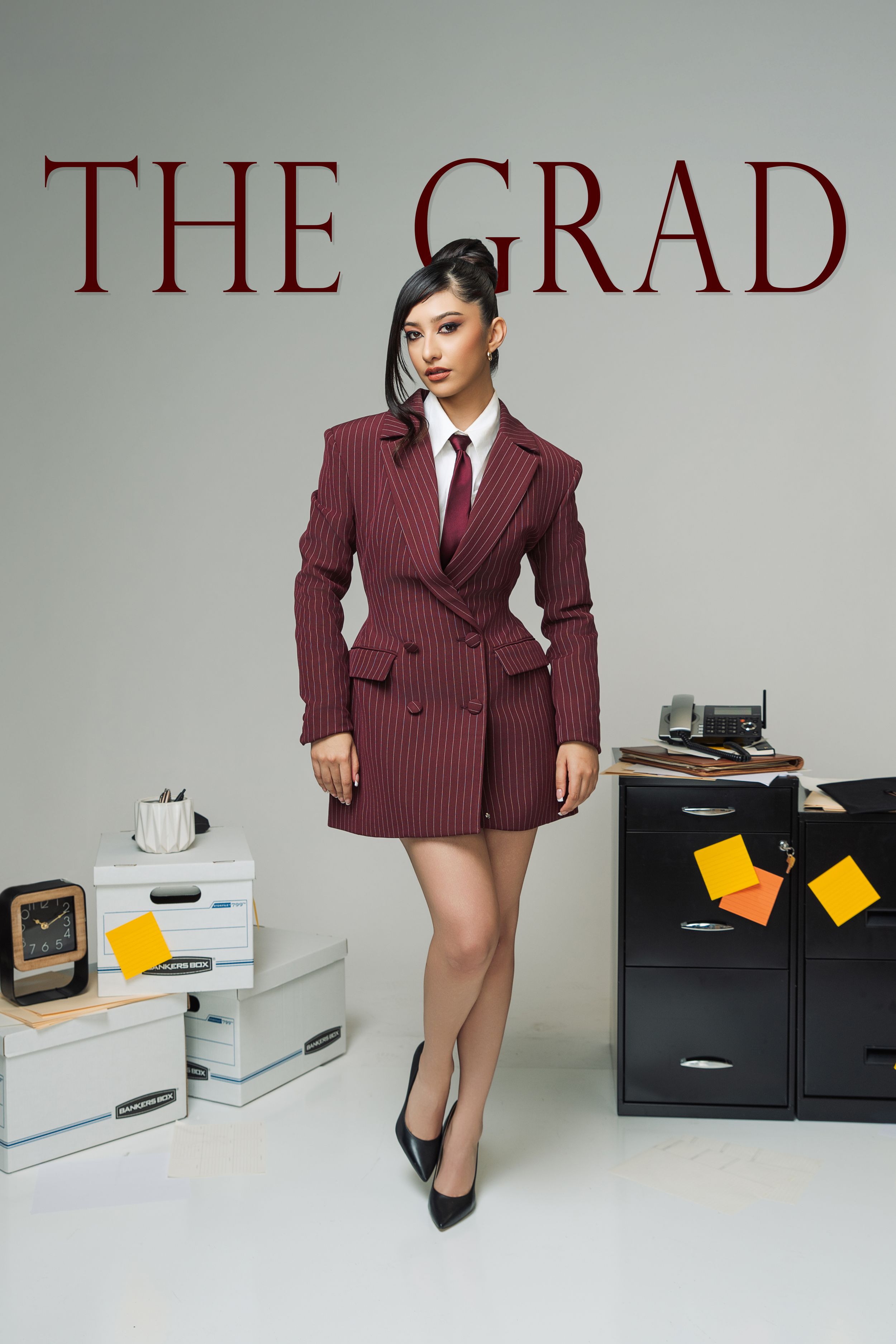 A woman in a burgundy pinstripe blazer dress and heels poses near office boxes and filing cabinet under 'The Grad' title.
