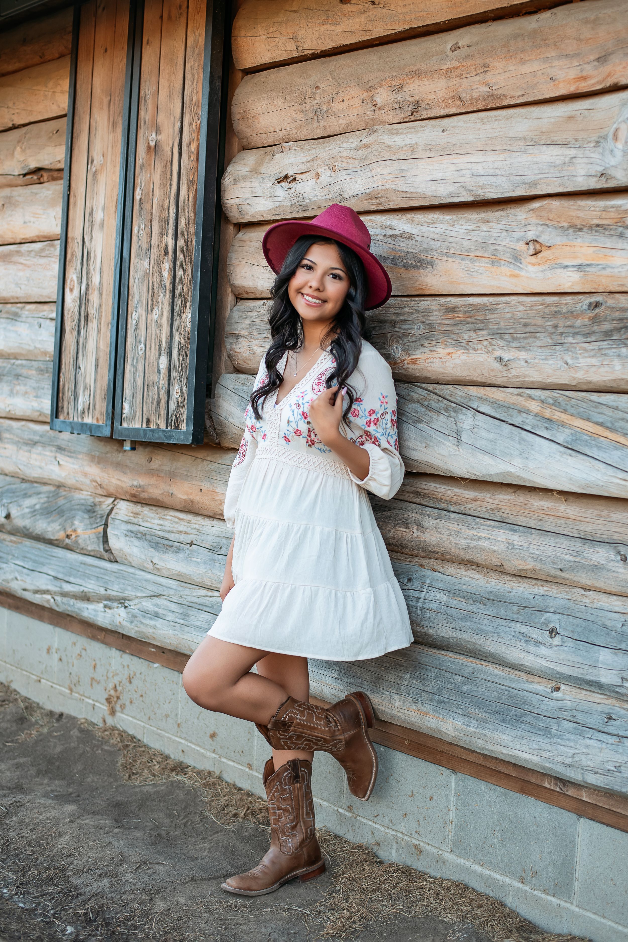 Country-style portrait in white dress and cowboy boots against rustic log cabin wall.