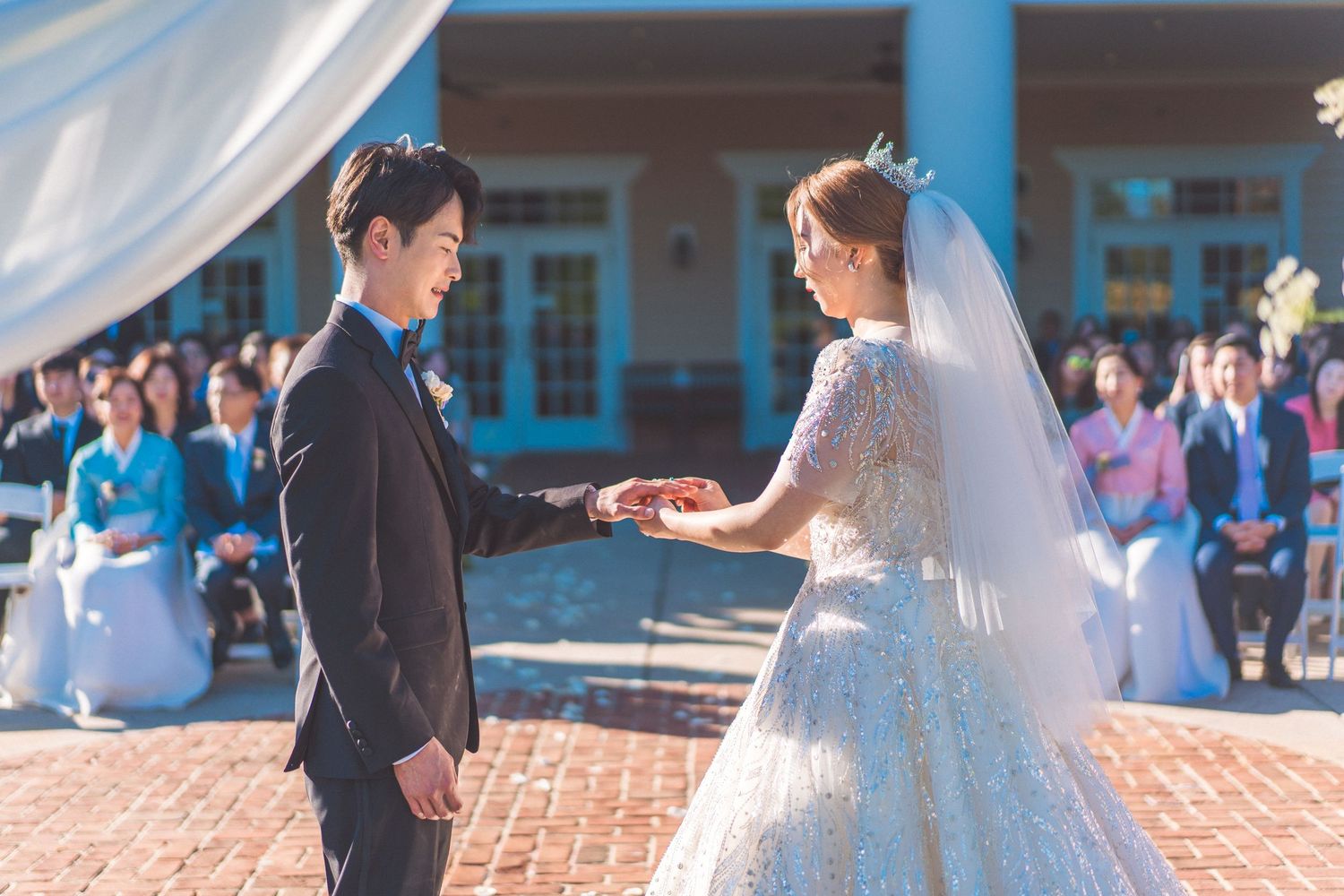 A romantic wedding ceremony exchange of rings on an outdoor brick patio with guests watching in the background.